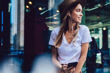 Moda kıyafetleri içinde, şapkalı, akıllı telefonlu, kulaklıklı, sokak kafesinde masaya yaslanmış ve gözlerini çevirmiş genç, çekici bir kadın.