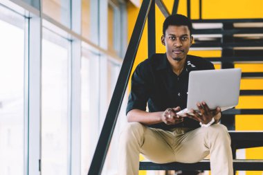 Kendine güvenen Afro-Amerikan erkek çalışan dizüstü bilgisayar ile ofis merdivenlerinde oturan ve kamera arıyor, resmi giyim koyu tenli kurumsal işçi coworking netbook üzerinde çalışıyor. Öğrenci adam