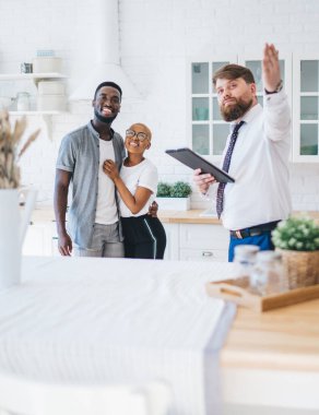 Profesyonel sakallı, resmi kıyafetli, pano takmış, modern güneşli dairede masaların yanında duran neşeli, kucaklayıcı etnik çifte mutfağını gösteren şişman bir adam.