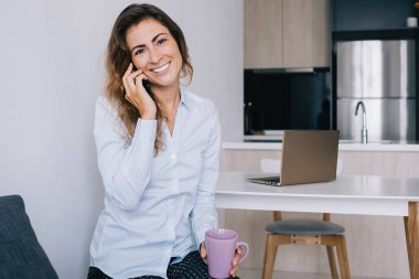 Kahverengi saçlı, bardakta içecekle oturan ve ofisteki kameraya bakıp gülümseyen çekici genç bayan.