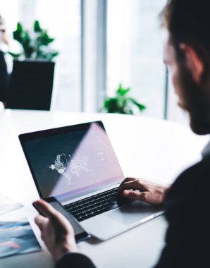 Resmi kıyafetli ve sakallı bir yönetici erkeğin, bilgisayarını kullanırken bilgisayarını kullanması, atölyede ticaret haritası üzerinde çalışması, bulanık kadın işçinin arka planında çalışması.