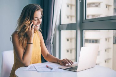 Sarı kolsuz bluz giyen, beyaz yuvarlak masada oturan, dizüstü bilgisayarla çalışan ve evde akıllı telefondan konuşan konuşkan genç bir kadın.