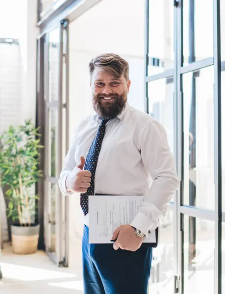Pleased bearded male worker in formal wear holding clipboard showing thumb up against African American couple in modern apartment happy with agreement