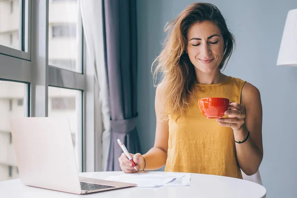 Thoughtful smiling young attractive woman in yellow sleeveless blouse sitting at white round table with laptop and red mug writing on paper at home
