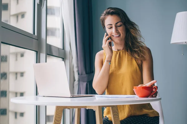 Happy young woman in yellow sleeveless blouse sitting at white round table with laptop and red mug communicating on smartphone at home