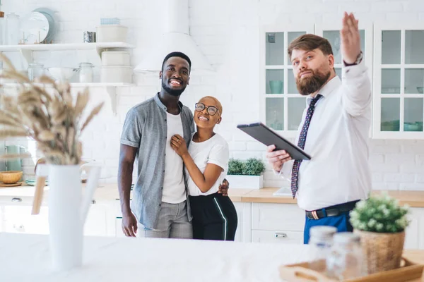 Professional bearded chubby man in formal outfit with clipboard demonstrating kitchen to cheerful embracing ethnic couple while standing next to table at modern sunny apartment