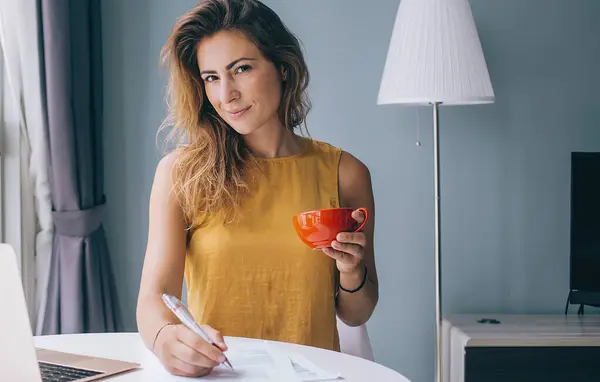Coquettish young attractive woman in yellow sleeveless blouse sitting at white round table with laptop and red mug writing on paper at home and looking at camera