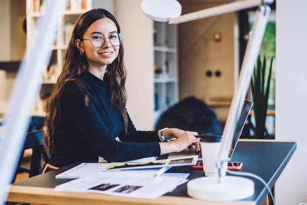 Smart casual female graphic designer in stylish optical eyewear sitting at coworking desktop with modern netbook for freelance work, portrait of Caucasian millennial woman blogger smiling at camera