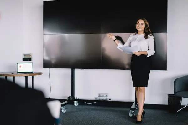 Young female in elegant outfit smiling and demonstrating presentation on blank screen for colleagues during business meeting in modern office