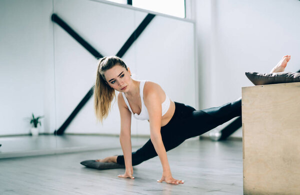 Serious fit confident sportswoman in black and white activewear focusing and stretching leg muscles leaning on wooden box and floor against sports resistance equipment and mirror wall in modern gym
