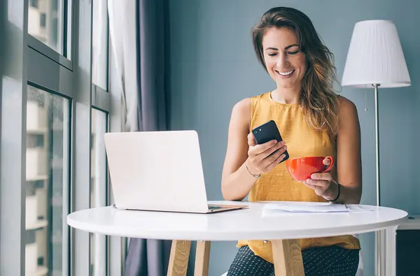 Young smiling woman sitting at round table with laptop while holding cup of tea and browsing Internet on smartphone in well-lit room