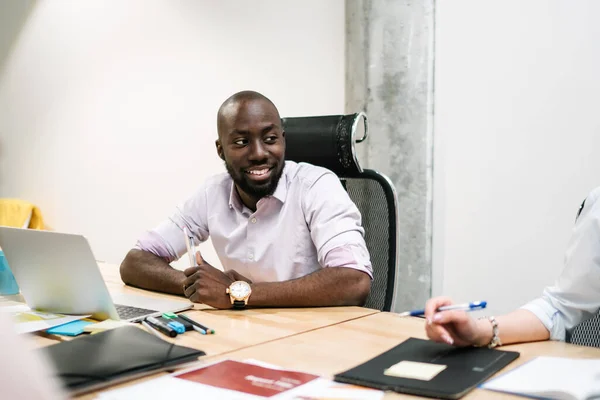 Cheerful smiling African American guy in casual clothes with watch creating cooperative startup looking at partner sitting at desk at workplace