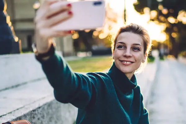 Cheerful young hipster girl with cellular phone sitting at historic urban setting and smiling at front camera for clicking selfie pictures and share to social networks for influence to followers