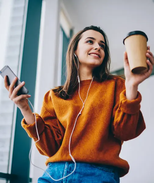Positive hipster girl smiling during listening motivation audio book via electronic headphones and online application for education holding coffee to go indoors, happy woman enjoying time for music