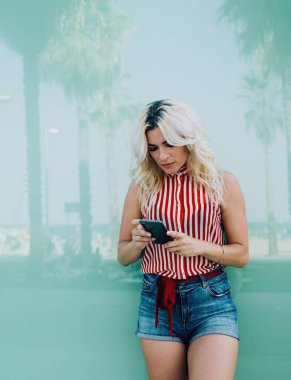 Günlük giysiler içinde, arkadaşlarıyla tropikal bir şehirde vakit geçirmek için telefon kullanarak sosyal medyada sohbet eden şehvetli bir kadın.