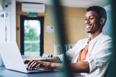 Gülümseyen, zeki, beyaz gömlekli, ofisteki masada oturmuş laptopunu karıştıran Afro-Amerikan adam.