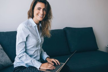 Modern yetişkin kadın dalgalı saçlar oturma odasında mavi koltukta oturuyor ve uzaktan modern dizüstü bilgisayarla çalışıyor kameraya gülümsüyor.