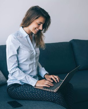 Çalışkan yetişkin bir kadın dizüstü bilgisayarla rahat bir koltukta oturuyor, gülümsüyor ve coşkuyla daktilo yazıyor.