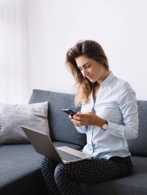 Tişörtlü modern bir kadın, oturma odasında dizlerinin üstüne çöküp koltukta otururken telefonla mesajlaşıyor.