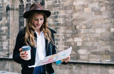 Güzel, modern kadın gezgin, şık bir şapka takıyor. Eski binanın yanındaki taş çitte oturuyor. Elinde tek kullanımlık bir fincan kahveyle şehir haritası okuyor.