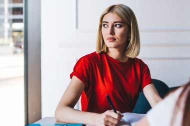 Düşünen, kırmızı tişörtlü sarışın kadın. Masada renkli kalemler ve çıkartmalarla notlar alıyor.