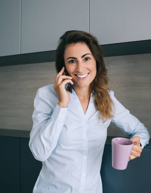 Gülümseyen serbest çalışan güzel bir bayan ya da mavi ofis gömlekli bir çalışan elinde fincanla durup akıllı telefonla konuşuyor ve kameraya bakıyor.