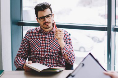 Gözlüklü, düşünceli, genç bir erkek çalışanın elinde defter var ve elinde dosyayla ofisteki bayan meslektaşıyla konuşuyor.