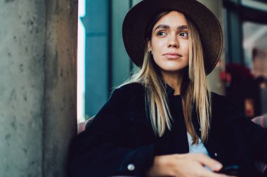 Şapkalı şık bir kadın, cep telefonu ve dizüstü bilgisayarla karanlık bir kafede rahat bir şekilde oturuyor.