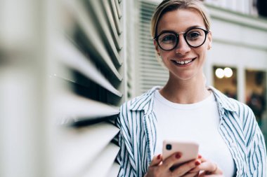 Neşeli sarışın kadın günlük kıyafetler giyiyor ve moda gözlükler takıyor. Gülümsüyor ve kameraya bakıyor. Duvara yaslanıyor ve akıllı telefonlara bakıyor.