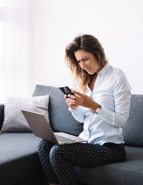 Oturma odasındaki kırışık suratlı dizüstü bilgisayarla koltukta oturmuş kafası karışık bir şekilde cep telefonu kullanan genç bir kadın.