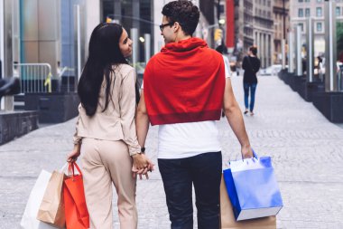 Gündüz vakti alışveriş torbalarıyla gezen modern, modaya uygun, çok ırklı, sevgi dolu genç bir kadın ve erkeğin arkasından.