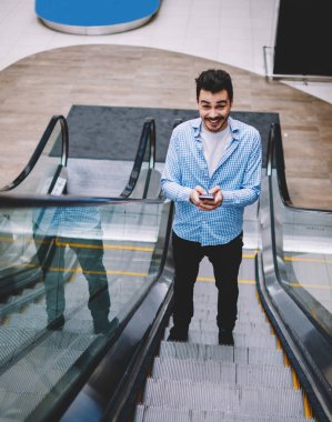 Elinde akıllı telefon gadget ile yürüyen merdiven üzerinde duran ve alışveriş merkezinde alışveriş merkezinde alışveriş merkezinde alışveriş yerini aramak için hücresel uygulama kullanarak zaman geçirirken gülümseyen neşeli hipster adam Portresi