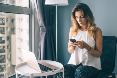 Üzücü odaklı, beyaz kolsuz bluz giyen esmer kadın yuvarlak masada oturmuş akıllı telefondan mesaj atıyor. Evde dizüstü bilgisayar kullanıyor.