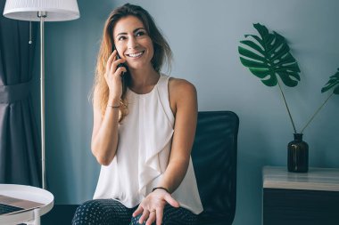 Sıradan giysiler içinde gülümseyen genç bir bayan bilgisayarla yan koltukta oturuyor ve akıllı telefondan konuşuyor ve eve bakıyor.