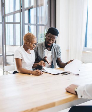 Düşünceli Afrikalı Amerikalı çift, emlakçının önünde masada oturmuş, emlak sözleşmelerini dikkatle okuyor ve tartışıyorlar.