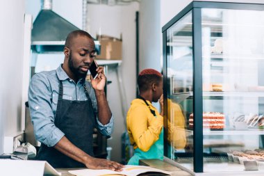 Afro-Amerikalı bir adam, hamur işi kafede çalışırken cep telefonuyla sohbet ediyor. Masanın arkasında elinde not defteriyle zenci bir kadın duruyor.