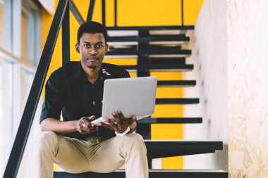 Kendine güvenen Afro-Amerikan erkek çalışan dizüstü bilgisayar ile ofis merdivenlerinde oturan ve kamera arıyor, resmi giyim koyu tenli kurumsal işçi coworking netbook üzerinde çalışıyor. Öğrenci adam