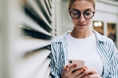 Gündelik kıyafetler giyen, şık gözlükler takan, akıllı telefon kullanan, duvara yaslanan ve son haberleri okuyan güzel bir bayan.