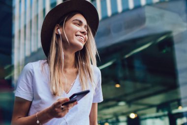 Şapkalı ve kulaklıklı mutlu bir kadın. Sıradan giysiler içinde. Sokakta duruyor.