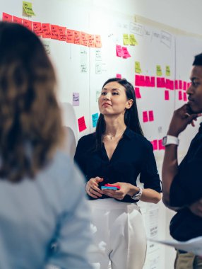 Çok ırklı erkek ve kadın iş arkadaşları resmi kıyafetler giyip renkli yapışkan notlarla hedefler belirliyor ve yaratıcı ofiste tartışıyorlar.