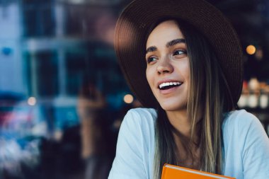 Keyifli ve neşeli genç bayan turuncu kitabı kucaklıyor ve kahvede pencerenin önünde otururken gülümsüyor ve başka tarafa bakıyor.