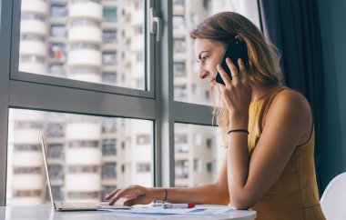 Sarı kolsuz bluz giyen, beyaz yuvarlak masada oturan, dizüstü bilgisayarda çalışan ve akıllı telefonuyla iletişim kuran ciddi bir kadının yan görüntüsü.