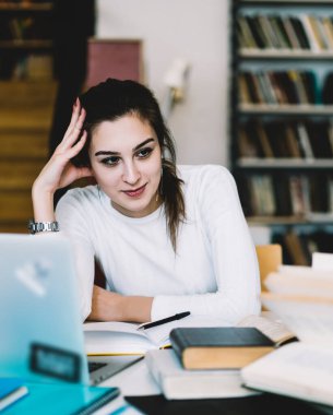 İçerik siyah saçlı beyaz ceketli bayan öğrenci masada oturuyor ve dizüstü bilgisayar ve kitaplarla ödev yaparken elini uzatıyor.