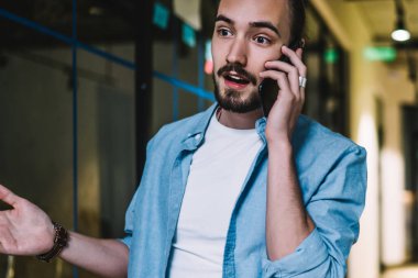 Gündelik giysiler içinde genç sakallı bir erkek akıllı telefondan konuşuyor ve ofis binasının koridorunda dikilirken haberlerle şaşırarak ellerini açıyor.