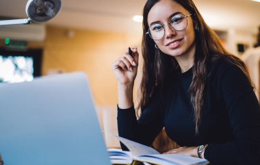 Moda gözlüklü akıllı kadın grafik tasarımcısı serbest çalışma için modern netbook ile birlikte çalışıyor, beyaz kadın blogcunun kameraya gülümseyen portresi