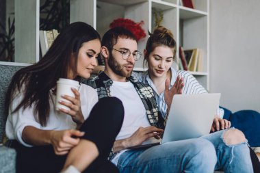 Genç sakallı adam dizlerinin üzerine çöküp siyah saçlı kadın ve plastik bardaklı genç bir kadınla film izliyor.