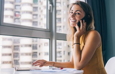 Sarı kolsuz bluzlu gülümseyen genç bir kadın beyaz yuvarlak bir masada oturuyor. Kağıt üzerinde çalışıyor ve kameraya bakarak akıllı telefondan iletişim kuruyor.