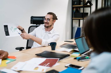 Dizüstü bilgisayarlarla dolu masada oturan, kahve ve klasörlerle dolu çok ırklı meslektaşlarına şema gösteren neşeli bir iş arkadaşı.