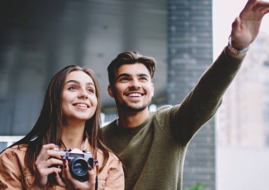 Aşık neşeli çift fotoğraf yapmak için vintage kamera kullanarak tarihte birlikte zaman harcama, mutlu yakışıklı adam smith kız arkadaşı için gösteren işaret ederken fotoğraf açık havada