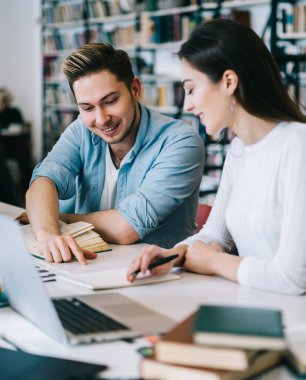 Çalışkan içerikli kadın ve erkek çalışırken gülüyor ve kütüphanede dizüstü bilgisayarlı açık kitaplarda bilgi arıyor.